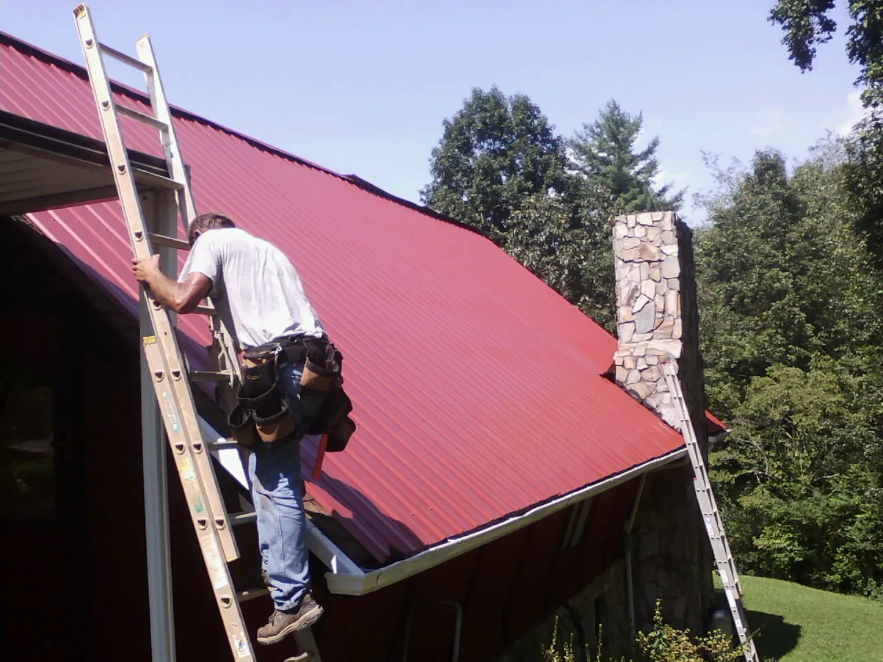 Roofer on a ladder beside a red metal roof and stone chimney — Soffit & Fascia Repair by Roofing Brothers in Pearblossom
